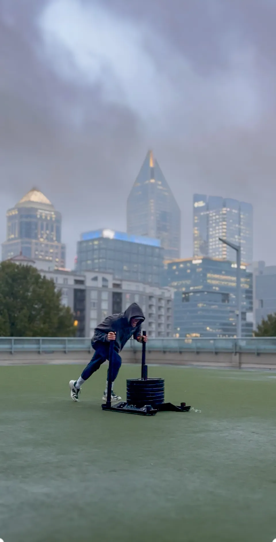 Thomas pushing a weighted sled on a rooftop turf with the Charlotte skyline behind